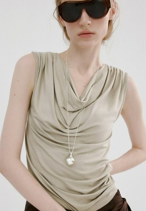 Femme portant de grandes lunettes de soleil noires, un haut beige sans manches drapé, un collier pendentif en argent, et un pantalon sombre, posant avec la main sur la hanche.