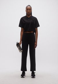 Γυναίκα που στέκεται, φορώντας αθλητικά ρούχα ολοκληρωτικά μαύρα, κρατώντας ένα διαφανές μπουκάλι νερού με μαύρο καπάκι, μπροστά σε ένα λευκό απλό φόντο.