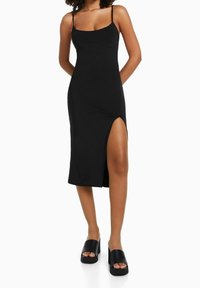 Robe midi noire à fines bretelles, corsage ajusté, fente latérale et texture lisse. Portée avec des sandales plateformes noires épaisses.