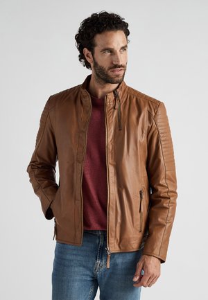 Veste en cuir marron avec un col montant, fermeture éclair sur le devant, épaules texturées et accents côtelés, assortie à une chemise rouge et un jean bleu.