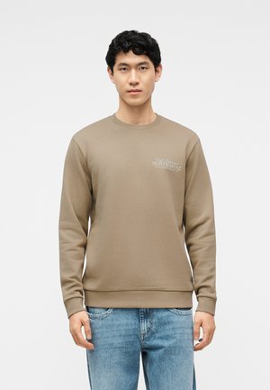 Νεαρός άνδρας που φοράει ανοιχτό καφέ φούτερ με το κείμενο "Montmartre" στο στήθος και ανοιχτόχρωμο μπλε τζιν, στέκεται μπροστά σε μονόχρωμο λευκό φόντο.