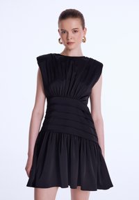 Robe noire sans manches avec bodice froncé, texture plissée et jupe évasée. Présente un tissu lisse et un design de taille structuré.