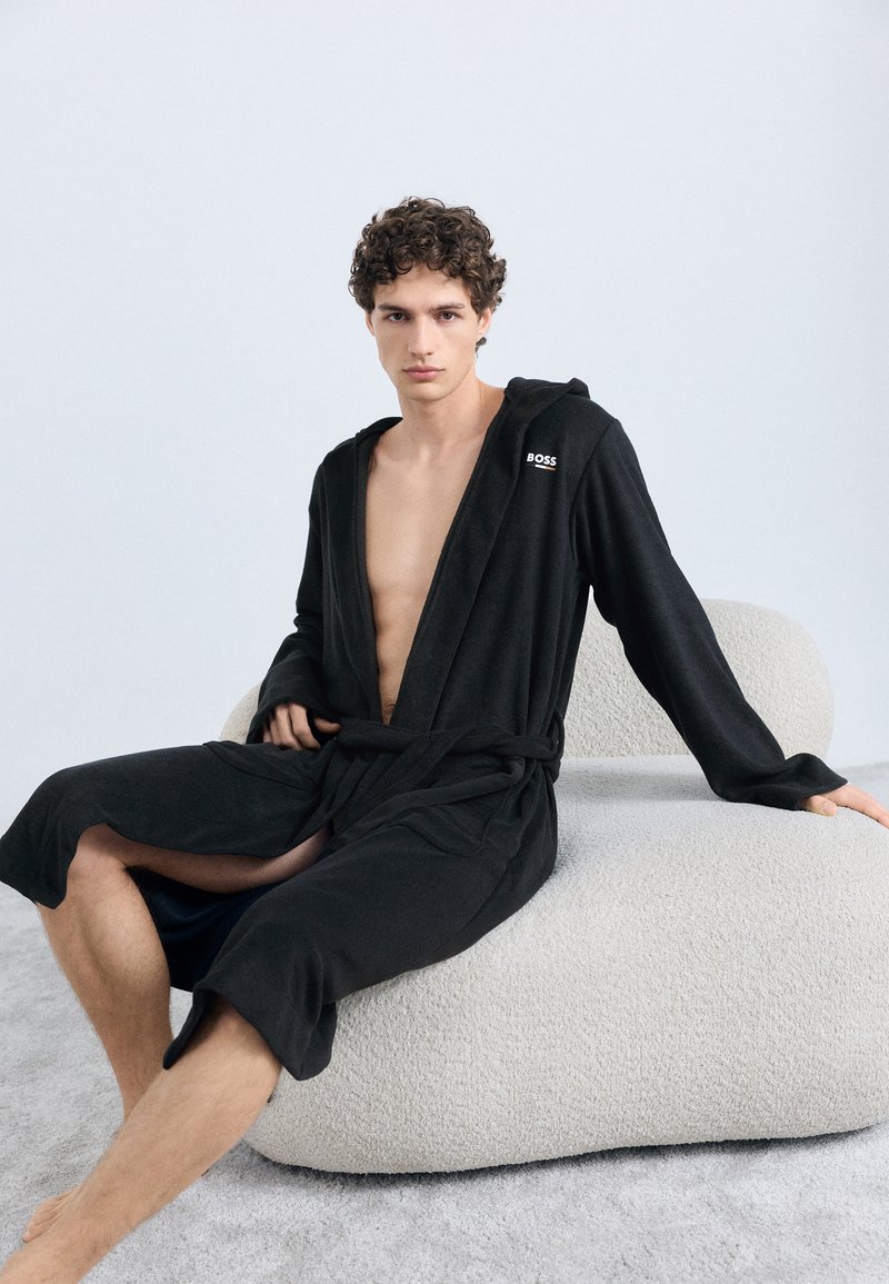 Robe de banho preta com capuz, cinto e logótipo no peito, feita de tecido macio. Modelo sentado numa cadeira texturada de cor clara.