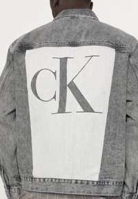 En person med kort hår bär en grå denimjacka som har en stor vit panel på ryggen med bokstäverna "CK" tryckta i mörkgrått.