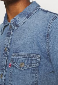 Světle modrá denimová košile s límečkem, patentkami a kapsou na prsu. Tkanina má jemnou strukturu a vybledlé oblasti.