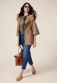 Chaqueta beige de gran tamaño con capucha, abierta sobre una blusa con estampado, combinada con jeans azul cortos, bailarina de color marrón claro y un bolso bicolor.