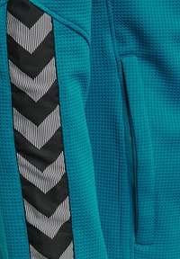 Tealfärgad textil med en nätliknande struktur, med en svart och vit zigzag-randig detalj längs sidan och kontrasterande sömnadsdetaljer.