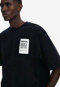 Black cotton t-shirt featuring a white patch with text "DESIGUAL DON'T BELIEVE EVERYTHING YOU THINK" and "BCN 1984." Relaxed fit with short sleeves.