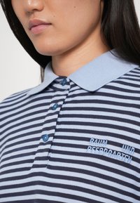 Prúžkovaná polokošeľa s námorníckymi a svetlomodrými vodorovnými pruhmi. Má svetlomodrý golier a gombíky a vyšívaný text na hrudi.