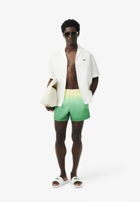 Camicia bianca a maniche corte su un torso nudo, abbinata a pantaloni da bagno verde sfumati, ciabatte bianche con logo e tiene una borsa chiara.