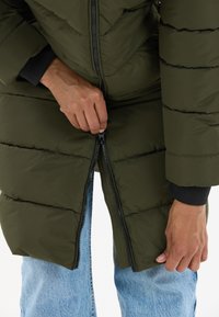 Olivengrøn pufferjakke med lynlås, med vandret quiltede sektioner og sorte manchetter. Modellen justerer lynlåsen nær blå jeans.