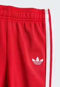 Røde treningsbukser med elastisk midje, hvite sidestriper og en liten hvit logo på nedre venstre ben. Glatt, lettvekt stoff.