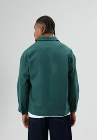 Grüne Fleecejacke mit langen Ärmeln, einem offenen Kragen und sichtbaren Steppnähten. Wird über einem weißen Hemd getragen, strukturierte Oberfläche.