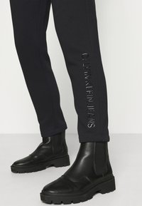 Svarta joggingsbyxor med tonal "CALVIN KLEIN JEANS" text på sidan. Kombination med svarta chelsea boots med tjock sula. Slät textur.
