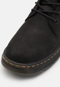 Bota de ante negra con puntera redonda, diseño de cordones negro, costuras amarillas en la suela y superficie superior texturizada.