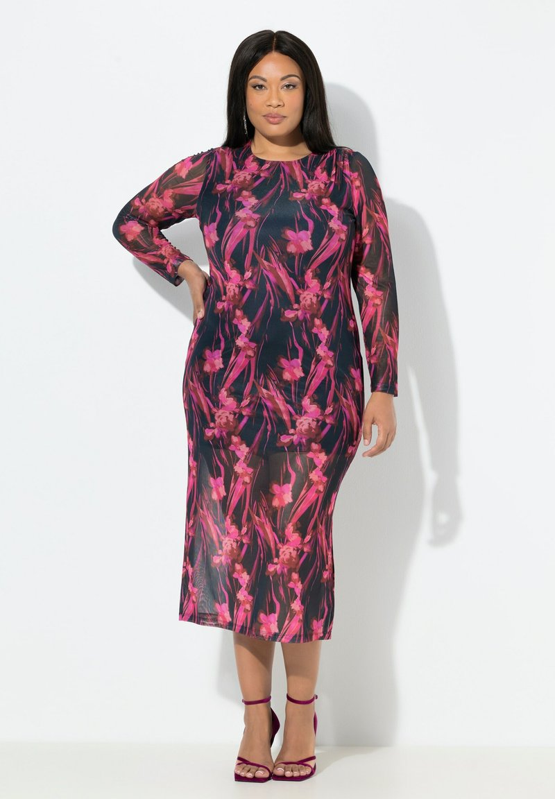 Vestido con estampado floral y mangas largas y transparentes; presenta flores rosas sobre un fondo oscuro; diseño hasta los tobillos; combinado con tacones fucsias.