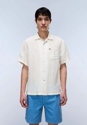 Junger Mann, der ein kurzärmeliges, elfenbeinfarbenes Hemd mit Knöpfen und leuchtend blaue Shorts trägt, steht vor einem schlichten hellen Hintergrund.