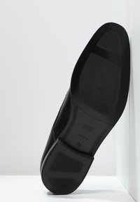 Chaussure en cuir noir avec une semelle en caoutchouc texturé, présentant des lignes de design subtiles et un marquage sur la semelle intérieure, incliné contre un fond blanc.