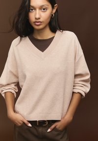 Unge kvinder med mørkt hår iført en beige V-hals sweater over en brun skjorte, brune bukser og guld hoop ørepropper, stående foran en brun baggrund.