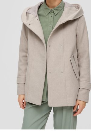 Persona con abrigo de lana con capucha de color beige claro sobre camisa verde salvia con botones y pantalones a juego, de pie frente a un fondo liso.