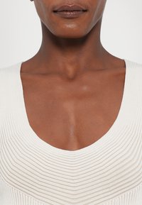 Gros plan sur le cou et le haut de la poitrine d’une personne portant un haut en maille gaufrée blanche avec un décolleté plongeant.