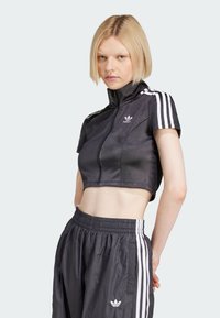 Zwarte cropped jas met een hoge kraag en een rits aan de voorkant, voorzien van witte drie-streep accenten op de mouwen, gecombineerd met zwarte sportbroek.