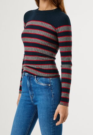 Kvinde iført en tætsiddende ribstrikket sweater med marineblå, grå og røde vandrette striber, kombineret med blå højttaljede jeans.