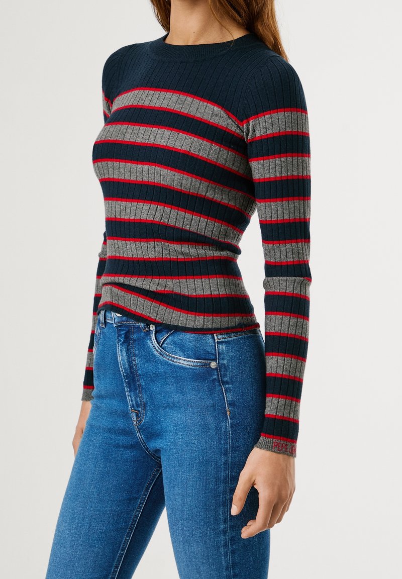 Femme portant un pull côtelé ajusté avec des rayures horizontales bleu marine, grises et rouges, associé à un jean bleu taille haute.