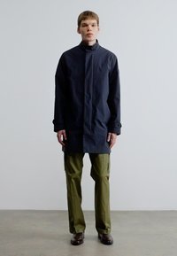 Tumesinine pikk mantel kõrge kraega, nööpidega ja küljetaskutega, kombineeritud oliivroheliste vabaaja pükstega ning pruunide kingadega lihtsal taustal.