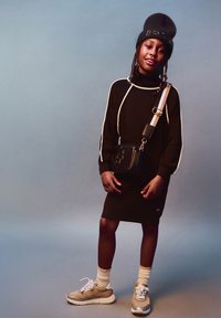 Robe noire en tricot avec des rayures blanches sur les côtés, portée avec un bonnet noir et un petit sac bandoulière noir. Des baskets beige et blanches complètent le look.