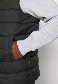 Czarna kamizelka puchowa z poziomymi przeszyciami, z boczną kieszenią i metką oznaczoną "EST 1993". Pod spodem widoczny rękaw szarego swetra.