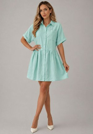 Femme portant une robe boutonnée à manches courtes vert menthe avec col et des talons hauts blancs, debout avec une main sur la hanche sur un fond uni.