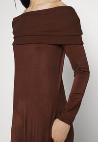 Brun off-shoulder klänning med ribbad halsringning, långa ärmar, sidofickor och mjukt, elastiskt tyg. Subtil lyster.