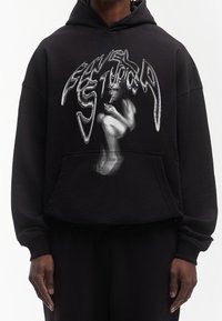 Sweat à capuche noir surdimensionné avec un design graphique déformé en blanc et gris. Le matériau semble doux avec une poche kangourou classique.