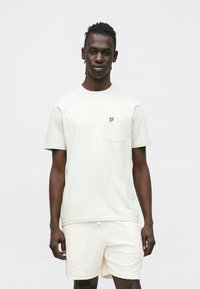 Light green t-shirt with short sleeves and a chest pocket featuring a small yellow logo. Paired with textured cream shorts.