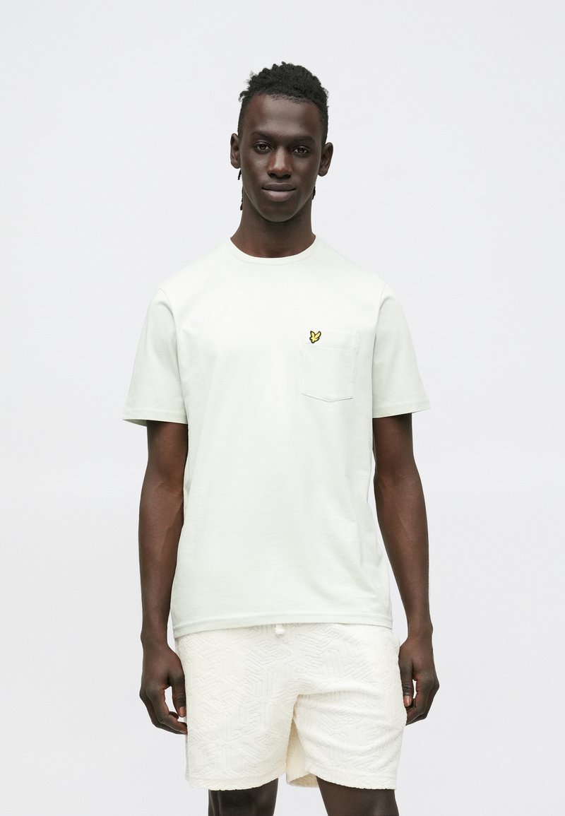 Light green t-shirt with short sleeves and a chest pocket featuring a small yellow logo. Paired with textured cream shorts.