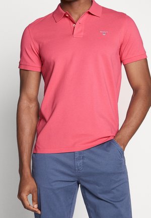 Homme portant un polo rouge à manches courtes avec col et un pantalon bleu texturé, une main dans la poche, sur fond uni.