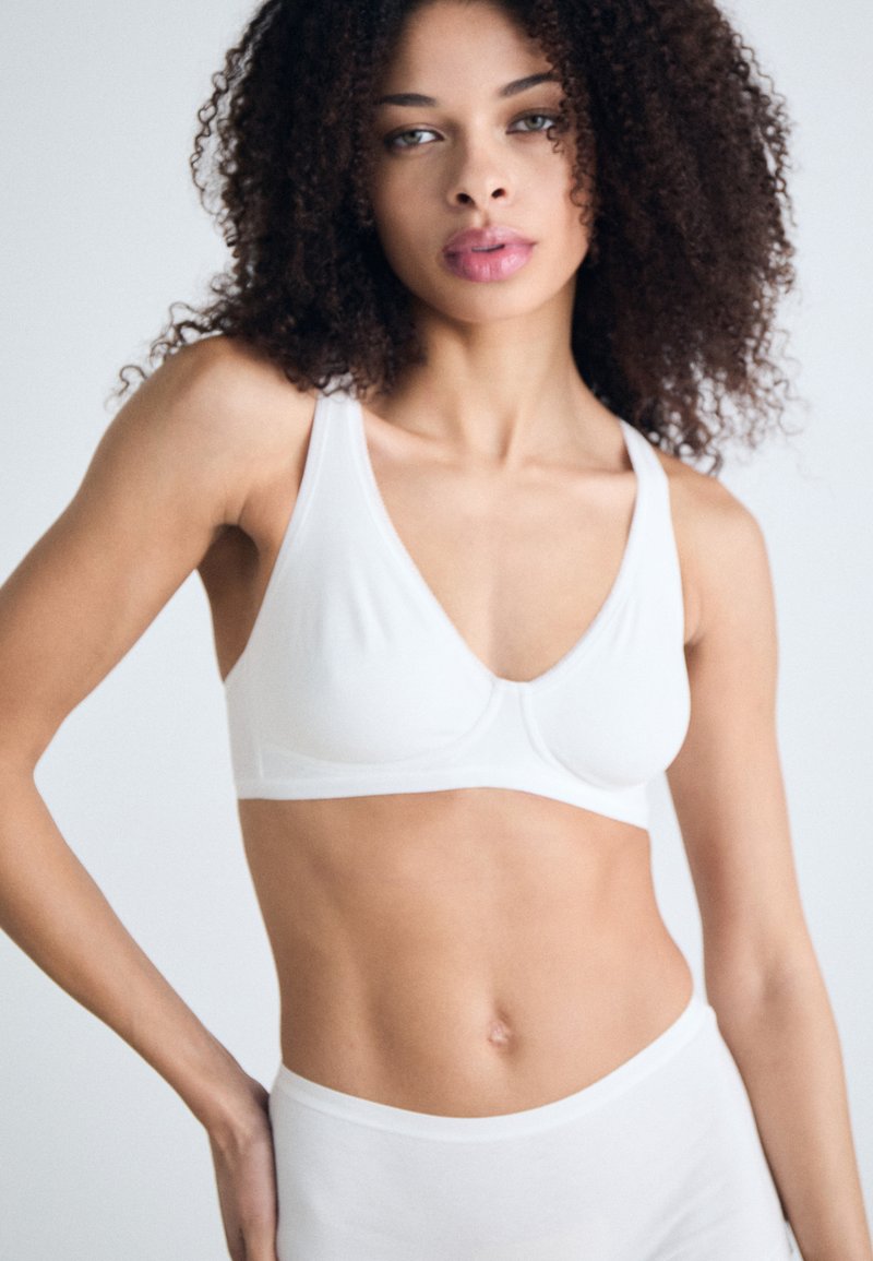 White wireless bra with a smooth texture, V-neck design, and wide straps. Model poses with mid-rise matching undergarments against a light background.