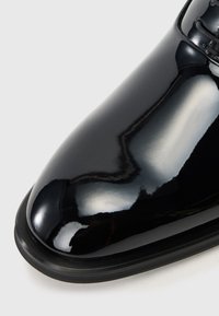 Μαύρο παπούτσι από πατεντέ δέρμα με γυαλιστερή επιφάνεια, κοφτή μύτη και ελάχιστη ραφή. Διαθέτει λεπτή καουτσούκ σόλα και σχέδιο με κορδόνια.