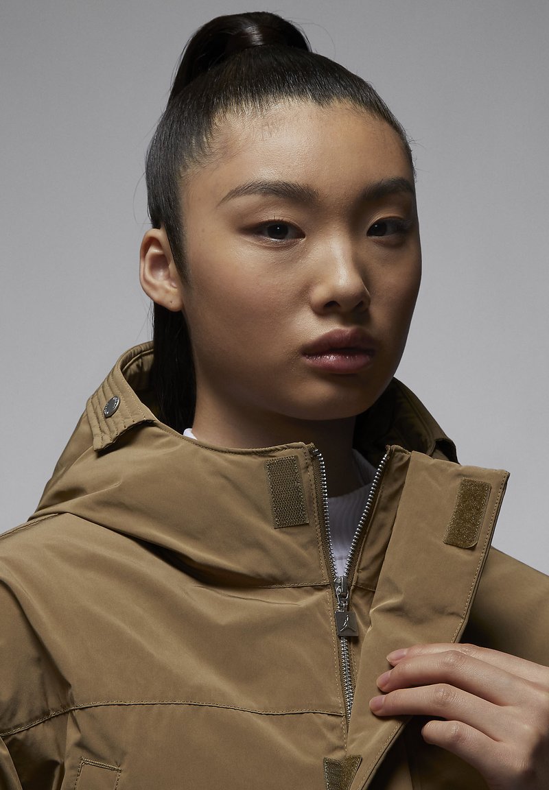 Chaqueta marrón con cuello alto y cierre de cremallera, con acentos de velcro y una textura suave y ligeramente brillante.