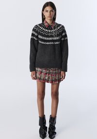 Maglione di carbone con motivi bianchi sul collo, abbinato a una gonna a quadri nei toni del rosso e del giallo, e stivali neri morbidi.