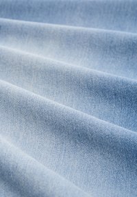 Light blue denim fabric with a textured weave, featuring subtle variations in shade across the surface and gentle folds.