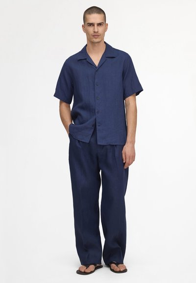 Homme portant une chemise bleu marine à manches courtes boutonnée, un pantalon ample assorti et des tongs noires, debout devant un fond blanc uni.