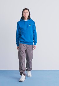Jaunas žmogus eina, vilkėdamas mėlyną Nike džemperį, pilkas krovinių kelnes ir baltus sportinius batelius, prieš paprastą šviesiai pilką foną.