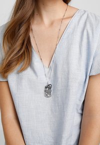 Femme portant un haut bleu clair texturé à col en V et un collier en chaîne argentée avec un pendentif en forme de cœur et une plaque militaire.