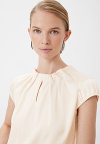 Light peach blouse with a gathered neckline and a small front keyhole. Short sleeves with subtle gathers, smooth fabric texture.