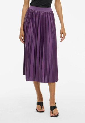 Femme portant une jupe midi plissée violette brillante et des sandales noires à talons, debout sur un fond blanc.