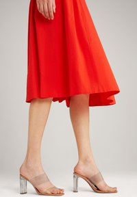 Robe midi plissée rouge avec un ourlet asymétrique. Une paire de mules à talons block transparents avec une lanière transparente sur le pied.