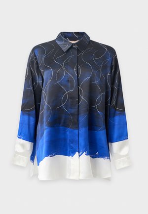 Femme aux longs cheveux noirs porte un jean bleu foncé ample, des chaussures blanches pointues et une chemise à manches longues noire, bleue et blanche à motifs, debout sur un fond blanc.