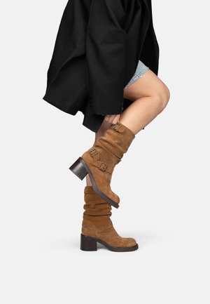Femme portant des bottes mi-mollet en daim couleur camel avec boucles et talons carrés, associées à un short en denim et un manteau noir.